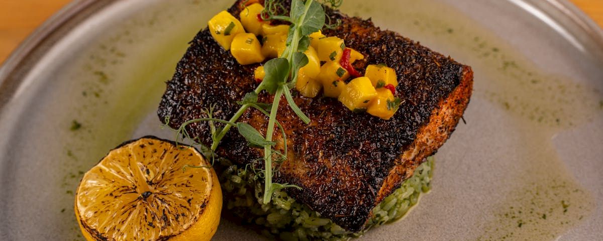A plated blackened fish fillet with mango salsa, rice/green garnish, and a lemon wedge.