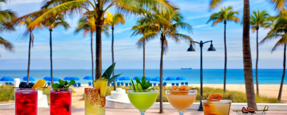 Colorful tropical cocktails with fruit garnishes sit on a table by a sandy beach, with palm trees swaying in the background.