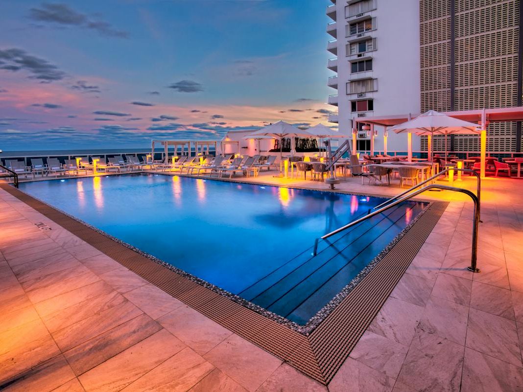 A resort hotel pool at sunset with lounge chairs, umbrellas, and colorful lights against tall buildings and the ocean horizon.
