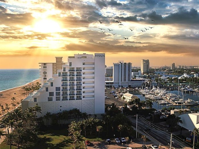 Sunset over a beachfront resort with palm trees, ocean on left, and a busy marina full of yachts on the right.