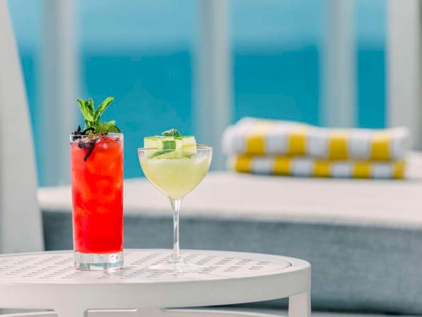 Two cocktails on a table: a tall red drink with mint and garnish, and a pale green cucumber-lemon cocktail in a glass.