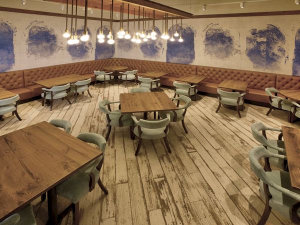 A modern lounge/caf&eacute; interior with wooden tables, upholstered banquette seating, pendant lights, and blue wall art murals.