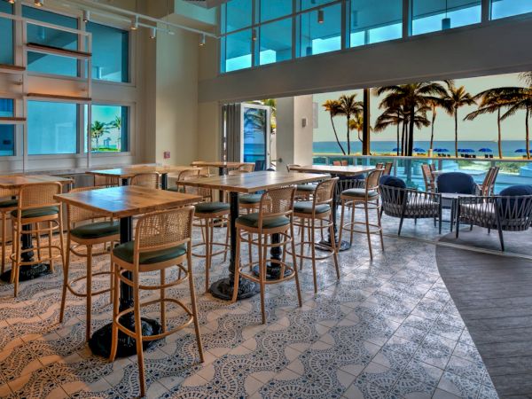 A modern beachfront caf&eacute; interior with wooden tables and bar stools, opening onto a patio with palm trees and ocean views.