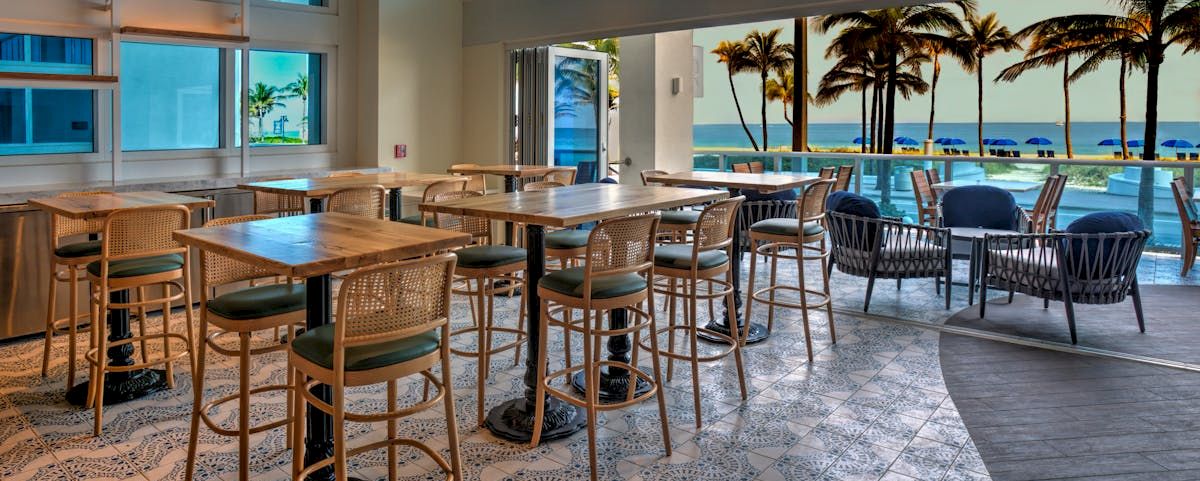 A bright hotel bar or lounge with wooden tables and chairs, opening to a tropical ocean view with palm trees and umbrellas outside.